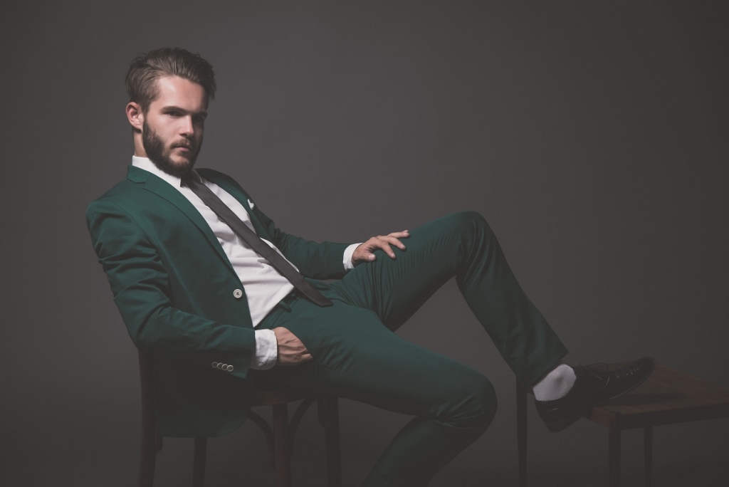 Business fashion man wearing green suit with white shirt black and tie. Sitting on wooden chair. Studio shot against grey.