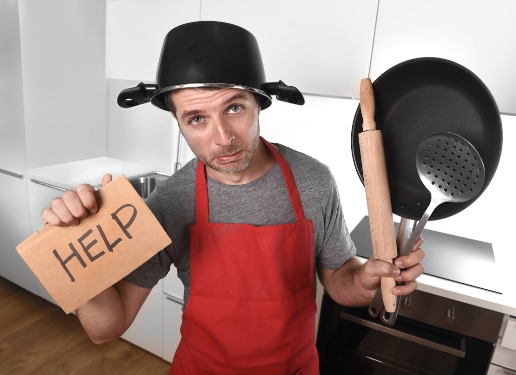funny 30s Caucasian man holding pan and household with pot on his head in red apron at home kitchen asking for help unable to cook showing panic on cooking with funny face expression