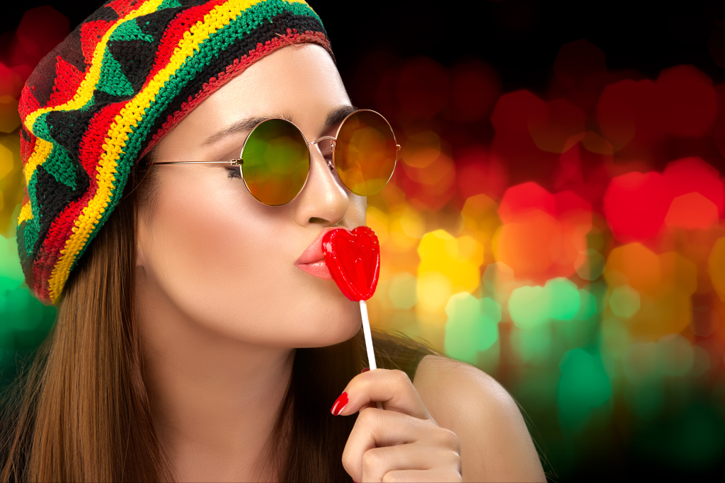 Stylish Party Girl in Rastafarian Hat and Trendy Round Sunglasses Kissing a Heart Shaped Lollipop. Close up portrait on an Colorful Abstract Background with Copy Space.