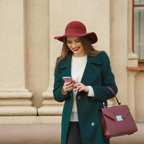 Fashionable Woman in a Bottle Green Coat and Burgundy Hat Smiling and Using Phone